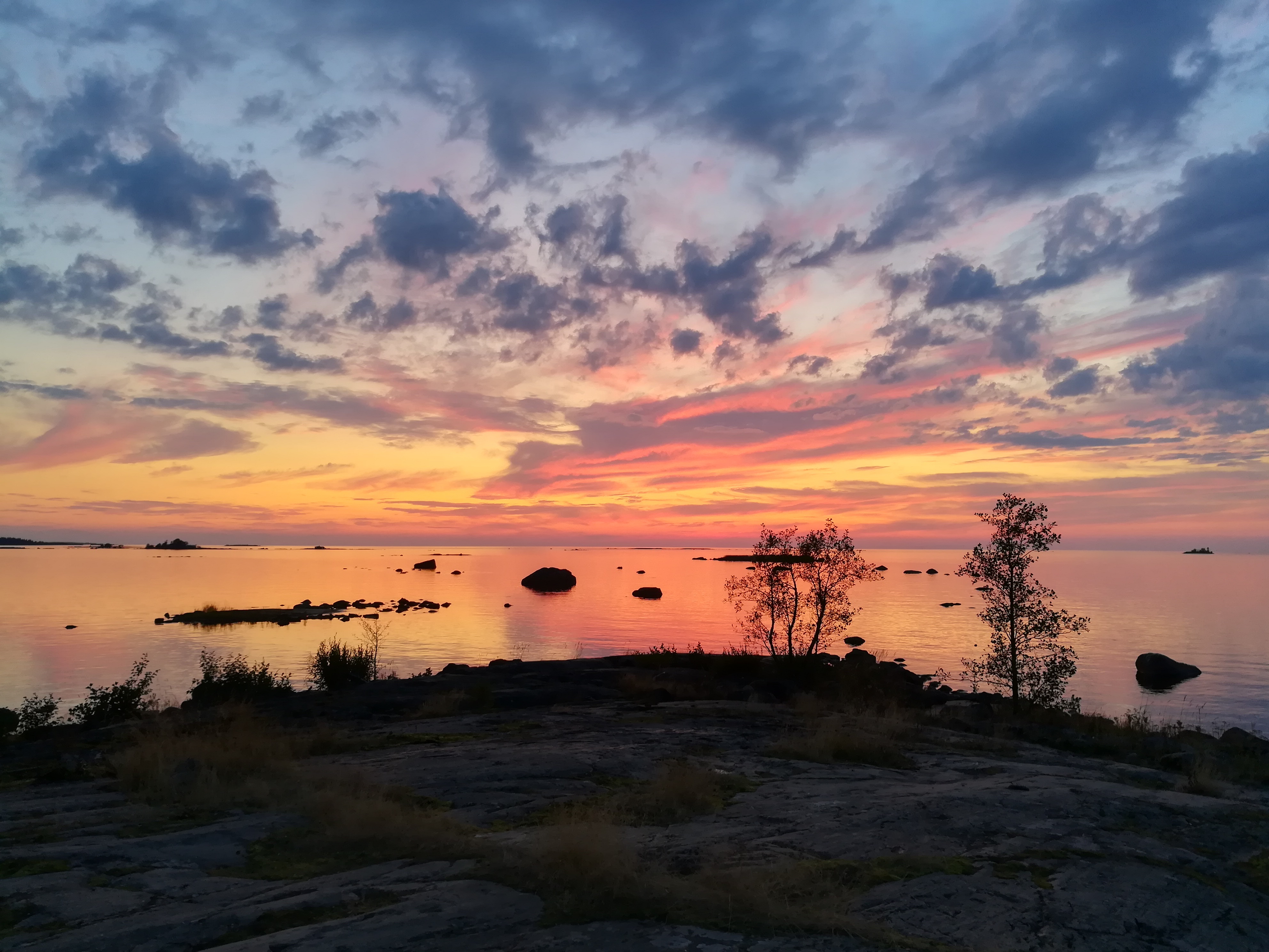 lilankeltainen taivas aurinko laskee mereen rannalla tummat kalliot ja pieniä puita
